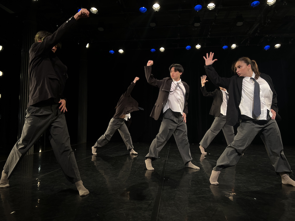 a company of young dancers male and female stand together in unison, legs in a wide stance, one hand on their hip the other in an upward motion as if to say 
